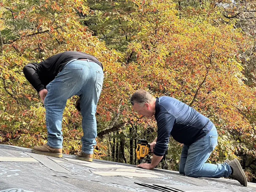 Two Ireland Contracting roofers working on a shingle roof with fall leaves behind them