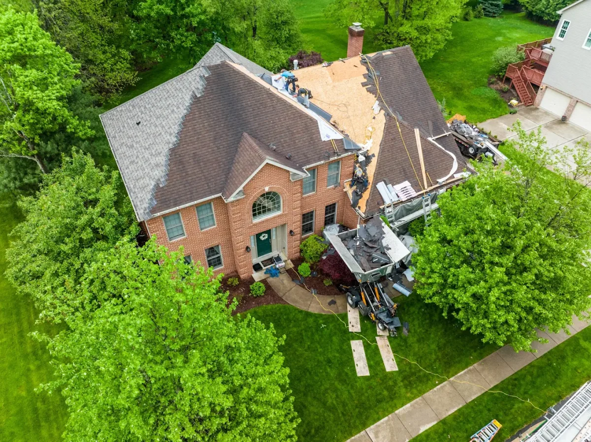 Aerial drone view of a brick home mid-roof-replacement with a crew tearing off to the decking and a debris trailer below